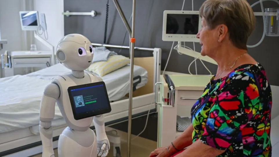 병원 병실에서 로봇이 침대에 앉아 있는 노인 여성과 대화하고 있다.