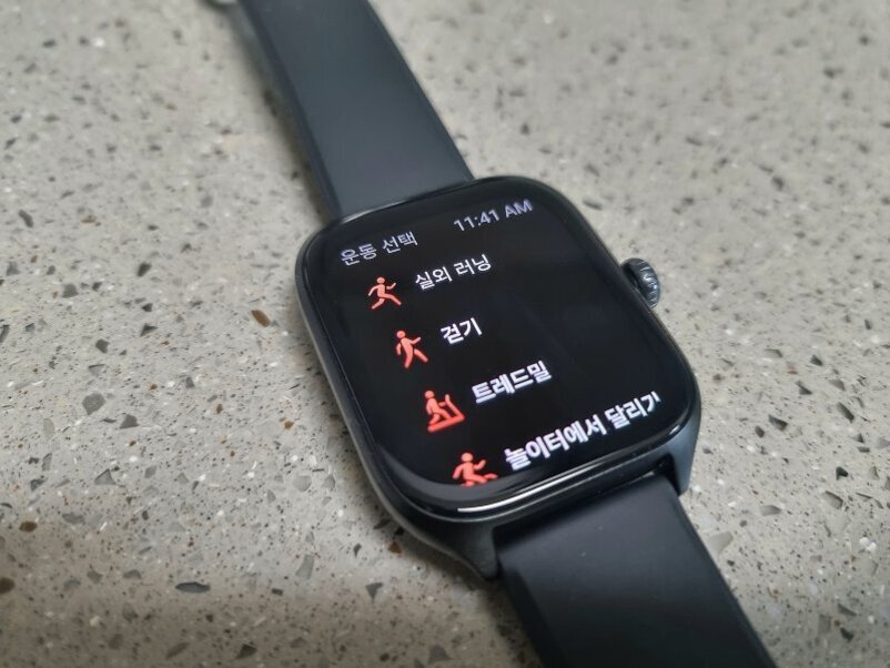 운동 선택 화면이 표시된 스마트워치. 실외 러닝, 걷기, 트레드밀, 놀이터에서 달리기 등 운동 종류가 한글로 나열되어 있다.