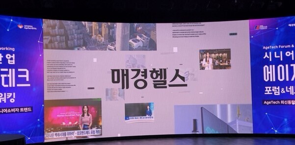대형 스크린에 '매경헬스'라는 글자가 중앙에 표시되어 있고, 양쪽에는 에이징테크 포럼&네트워킹 관련 안내 배너와 다양한 뉴스 이미지가 보인다.
