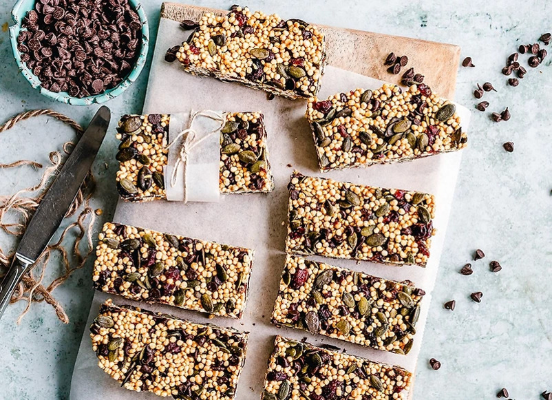 여러 개의 곡물과 씨앗, 건과일이 들어간 그래놀라 바가 도마 위에 가지런히 놓여 있다. 옆에는 초콜릿 칩이 담긴 그릇과 나이프가 보인다.