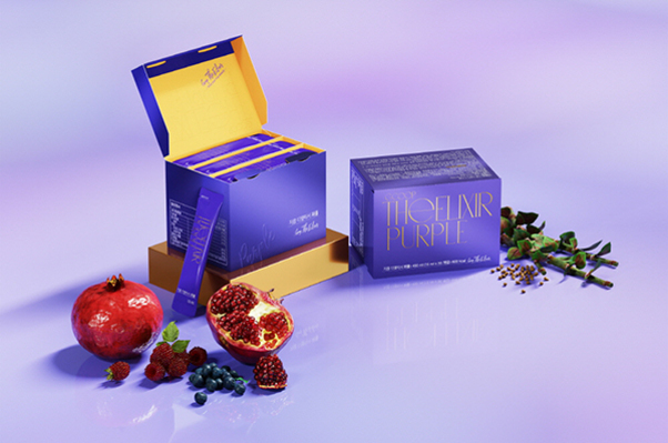 보라색과 노란색 포장의 건강기능식품 박스와 스틱, 주변에는 석류, 블루베리, 허브 등이 장식되어 있다.