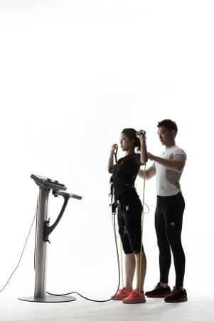 여성이 EMS 트레이닝 슈트를 입고 운동을 하고 있으며, 남성 트레이너가 그녀를 보조하고 있다. 앞에는 전자 장치가 있다.