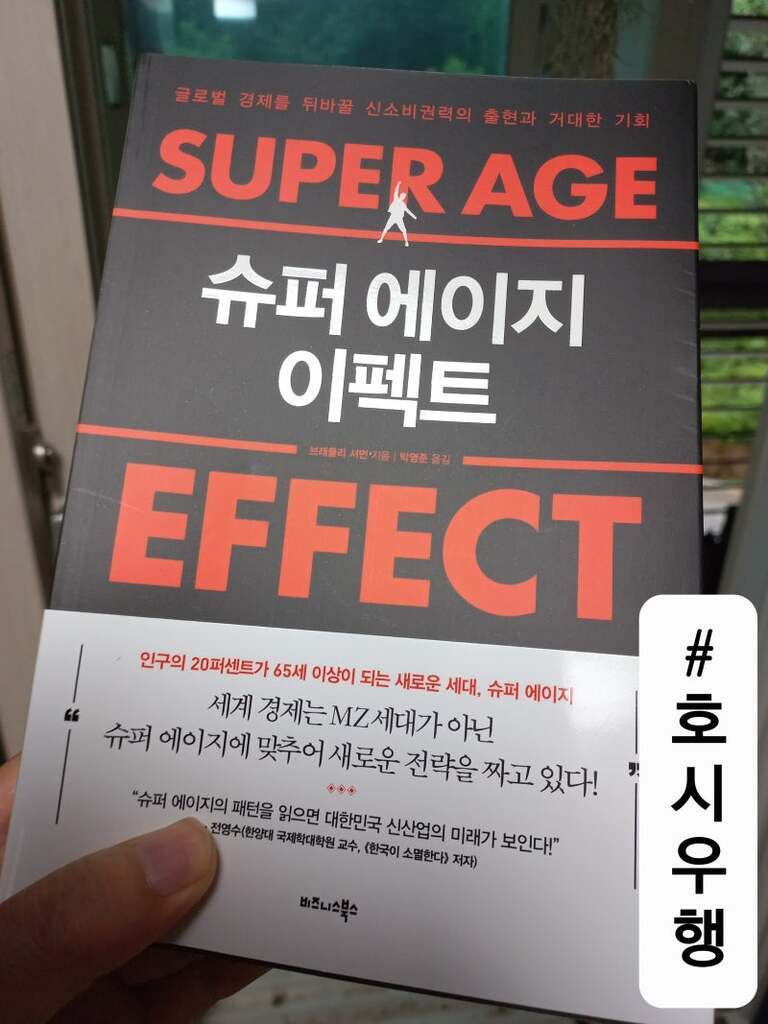 사람이 'SUPER AGE 슈퍼 에이지 이펙트'라는 제목의 책을 들고 있는 모습. 책 표지에는 고령화 사회와 관련된 메시지가 적혀 있으며, 하단에는 '#호시우행'이라는 해시태그가 있는 흰색 띠지가 붙어 있다.
