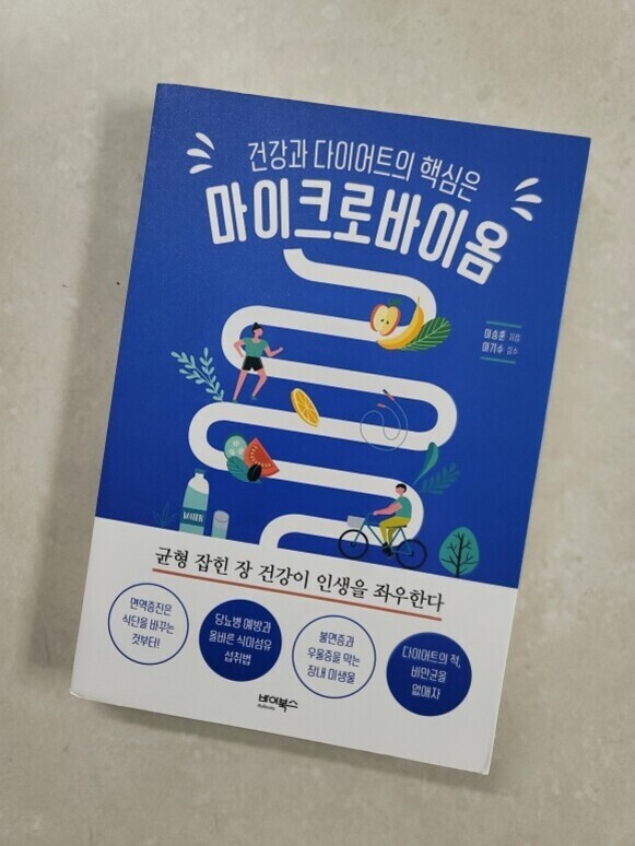 '건강과 다이어트의 핵심은 마이크로바이옴'이라는 제목의 한국어 책 표지. 파란색 배경에 장 모양을 형상화한 흰색 곡선 위로 운동하는 사람, 과일, 채소 등이 그려져 있다. 하단에는 '균형 잡힌 장 건강이 인생을 좌우한다'라는 문구와 여러 관련 정보가 원 안에 담겨 있다.