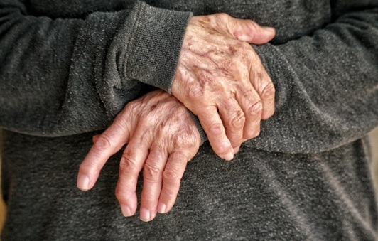 회색 옷을 입은 사람이 두 손을 겹쳐서 앞으로 모으고 있는 모습. 손에는 주름과 반점이 있어 노인의 손으로 보인다.