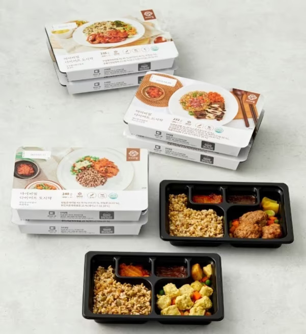 여러 개의 다이어트 도시락이 포장된 채로 쌓여 있고, 앞쪽에는 포장을 벗긴 상태의 도시락 두 개가 보인다. 각 도시락에는 밥, 반찬, 소스 등이 칸칸이 나뉘어 담겨 있다.