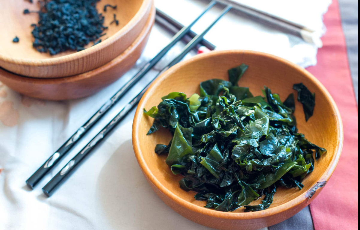 나무 그릇에 담긴 삶은 미역과 뒤쪽에는 잘게 썬 마른 미역, 젓가락이 놓여 있는 모습