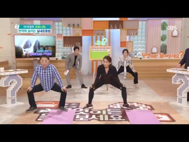 네 명의 사람들이 실내 스튜디오에서 운동 자세를 따라하며 스트레칭을 하고 있다. 바닥에는 운동 매트와 안내 문구가 있으며, 뒤쪽에는 TV 화면과 여러 소품들이 배치되어 있다.