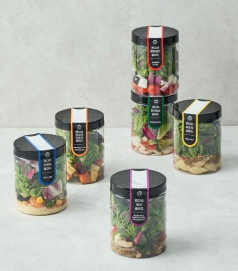 다섯 개의 투명 플라스틱 용기에 다양한 샐러드가 담겨 있으며, 각 용기에는 색깔별 라벨이 붙어 있다. 배경은 밝고 심플한 콘크리트 느낌의 표면이다.
