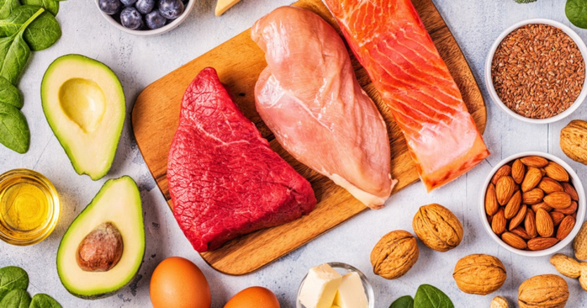 나무 도마 위에 소고기, 닭가슴살, 연어가 놓여 있고 주변에 아보카도, 시금치, 블루베리, 달걀, 견과류(아몬드와 호두), 식용유, 버터와 씨앗 등이 진열되어 있는 건강식 재료들.