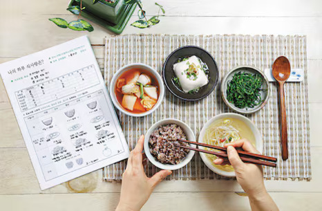 밥, 국, 반찬(두부, 나물, 김치)이 담긴 그릇들이 식탁에 놓여 있고, 한 사람이 젓가락으로 밥을 먹으려는 모습. 옆에는 식단표가 있다.