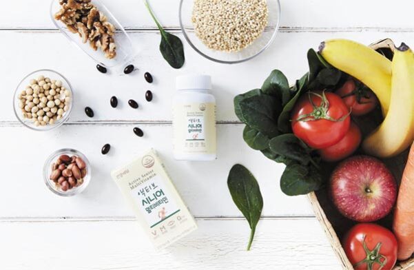 흰색 테이블 위에 각종 채소와 과일(바나나, 시금치, 토마토, 사과, 당근), 견과류(호두), 콩류(검은콩, 대두), 곡물 한 그릇, 건강기능식품 박스와 병이 놓여 있다.